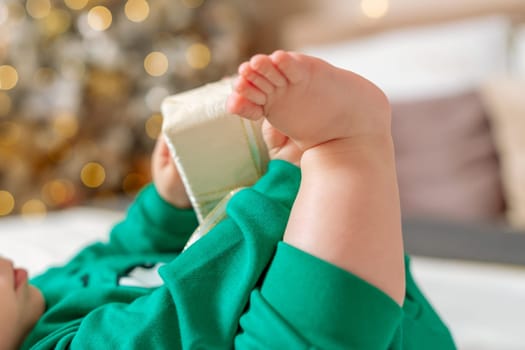 Kid gift. A little boy in a green suit holds a gift box with his feet and hands in bed