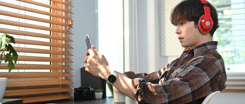 Handsome asian man in stylish casual wear listening to music in headphone and surfing internet on digital tablet.