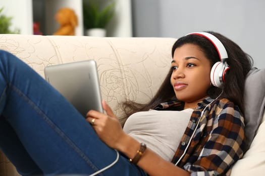 Happy african american young woman lying on sofa with tablet computer and headphones listening to music at home. Applications for listening to music watching movies