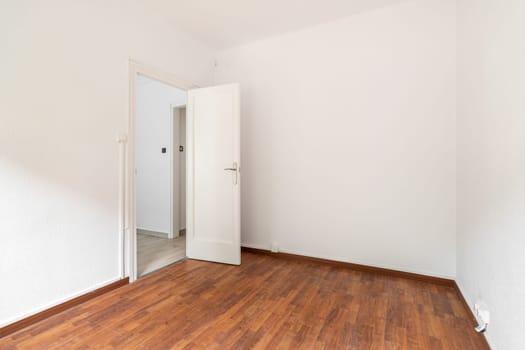 Empty spacious room well lit by daylight. The floor is dark brown wood parquet. White walls with a doorway opening onto a corridor. Through the open door you can see part of the rest of the apartment