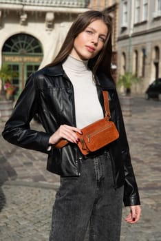 young brunette posing outside in jeans, white sweater and leather jacket with a small orange belt bag