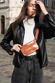 young brunette posing outside in jeans, white sweater and leather jacket with a small orange belt bag