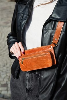 woman posing outside in jeans, white sweater and leather jacket with a small orange belt bag