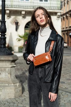 young brunette posing outside in jeans, white sweater and leather jacket with a small orange belt bag