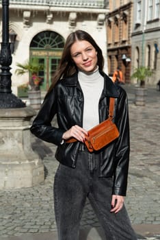 young brunette posing outside in jeans, white sweater and leather jacket with a small orange belt bag