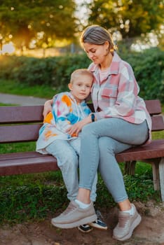 mother and son sit on a park bench in the rays of the setting sun. the concept of a family. Mother's Day. beautiful girl (mother) with a boy (son) in the park in the park are sitting on a bench at sunset.