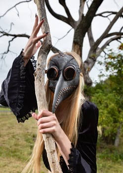 Natures beauty with a touch of mystery. A mystical and captivating image - a blonde woman in a crow mask posing near a withered tree in nature, creating an aura of mystery and beauty.