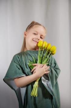 Portrait of a little blonde girl with bouquet of spring yellow flowers on a light background. Child in green dress holding a bouquet of tulips in hands. Spring concept