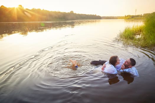 Beautiful adult couple having hugs and fun on nature in the water of a river or lake in the summer evening at sunset. A guy and a girl swim and relax outdoors in clothes in white shirts and jeans