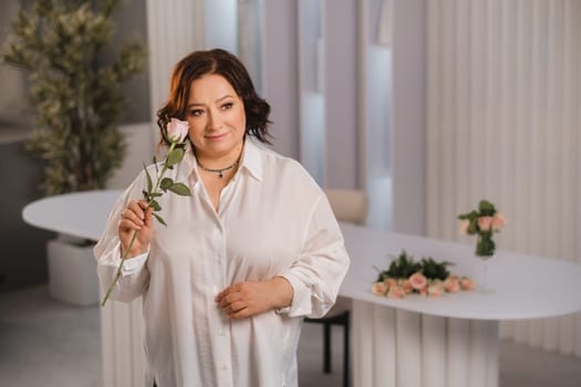 An adult woman in a white shirt holds one rose in her hands while standing in the interior. Pink roses.