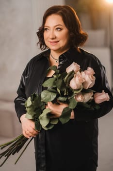 a stylish adult woman in black leather clothes stands with a bouquet of pink roses in the interior.