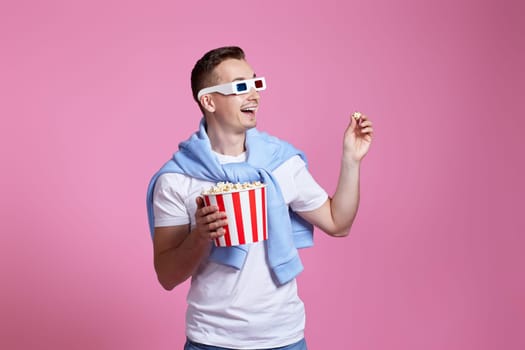 handsome man in 3d glasses, white t-shirt showing popcorn and looking at the side on pink background