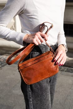 young brunette posing outside in jeans, white sweater with a small orange handbag