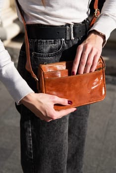 young brunette posing outside in jeans, white sweater with a small orange handbag