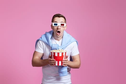 scared handsome man in 3d glasses holding bucket of popcorn on pink background