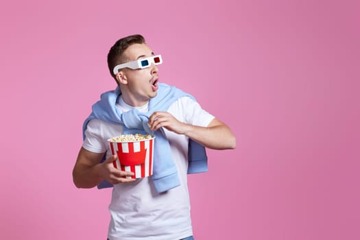 scared handsome man in 3d glasses holding bucket of popcorn on pink background