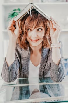 Woman book on her head. Surprised happy brunette in a jacket with a book on her head looks away against the backdrop of a bright office