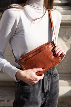 young brunette posing outside in jeans, white sweater with a small orange handbag