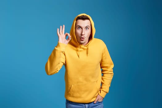 happy young man in yellow sweatshirt showing Ok gesture isolated on blue background.