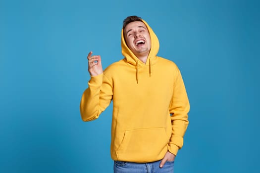 positive handsome man in yellow sweatshirt laughing on blue background