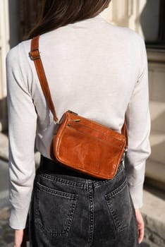 young brunette posing outside in jeans, white sweater with a small orange handbag