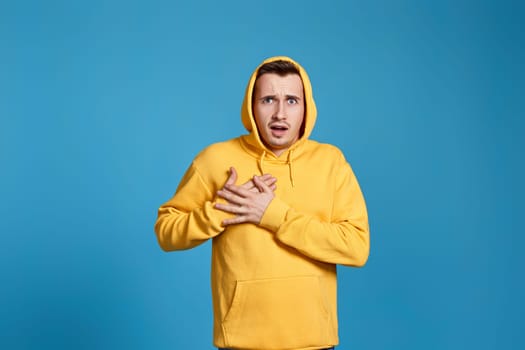 young man in yellow sweatshirt with hands on chest with closed eyes isolated on blue background.