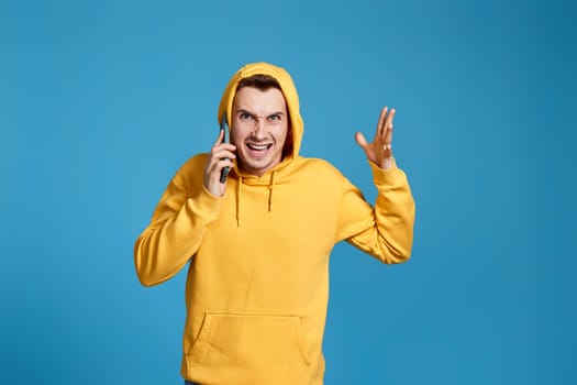 Worried nervous man in yellow sweatshirt holding mobile phone and feel outraged isolated on blue background