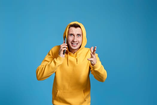 Upset angry man in yellow sweatshirt holding mobile phone and feel outraged isolated on blue background. Fear and upset for mistake.