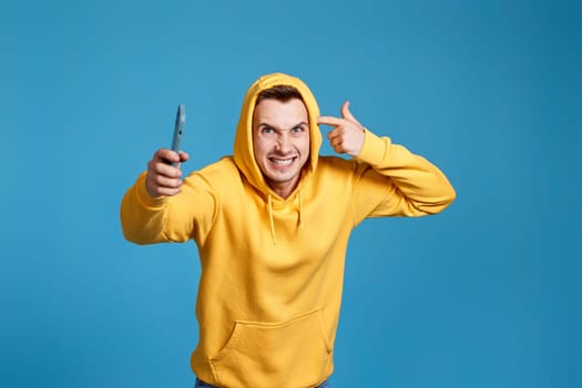 angry and frustrated man in yellow sweatshirt holding mobile phone and feel outraged isolated on blue background
