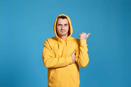 frowning man in yellow sweatshirt pointing at copy space isolated on blue background
