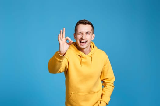 happy young man in yellow sweatshirt winking and showing Ok gesture isolated on blue background.
