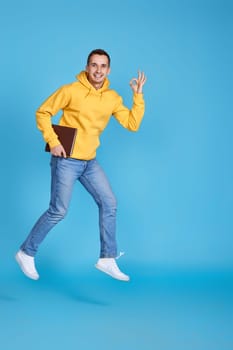 jumping very excited man with book showing Ok gesture on blue background. Full length