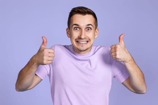 positive man showing thumbs up and looking at camera on pastel purple studio background