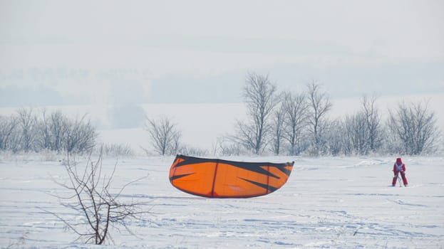 A girl in a red jumpsuit, snowkiting, parachuting in the field in winter. Lying kiting. Girl is kiting in the field.