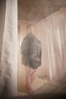 Portrait of sexual body positive adult lady woman in intimate in tender space indoors in a bedroom. Plump girl poses in black suit jacket during boudoir style photo shoot