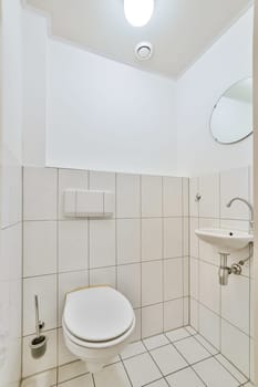 a small bathroom with white tiles on the walls and floor, there is a round mirror above the toilet bowl