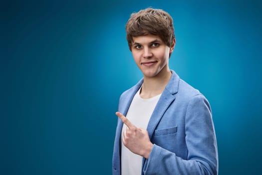 happy caucasian young man in jacket pointing finger at copy space on blue background