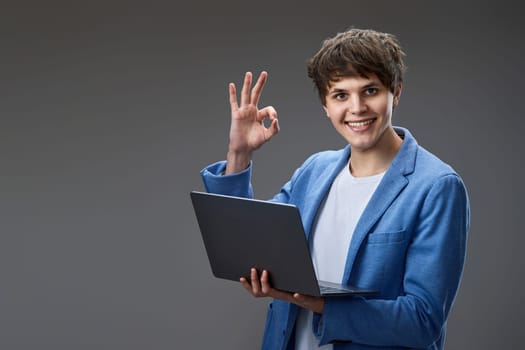 Successful caucasian young man using laptop computer watching webinars and showing Ok gesture isolated on gray background