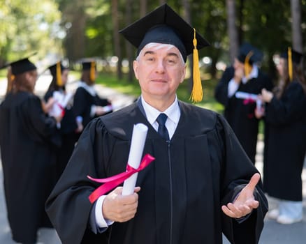 A group of graduates in robes outdoors. An elderly student rejoices at receiving a diploma