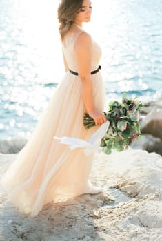 Bride with a bouquet stands with her head turned on a rocky seashore. High quality photo