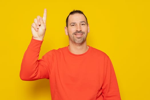 Handsome hispanic man with a beard in a red sweatshirt raising his index finger excited, he has had a great idea and he wants to tell everyone about it. Isolated on yellow background