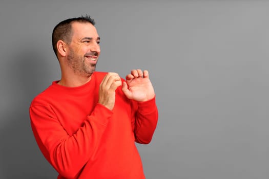 Hispanic man in his 40s wearing a red sweater poses embarrassed to the side with his hands in a defensive position, isolated on gray background
