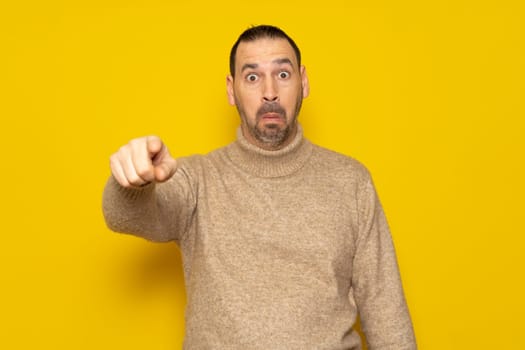 Bearded hispanic man in his 40s wearing a beige turtleneck pointing at something scary, he is very scared and feeling totally terrified. Isolated on yellow background