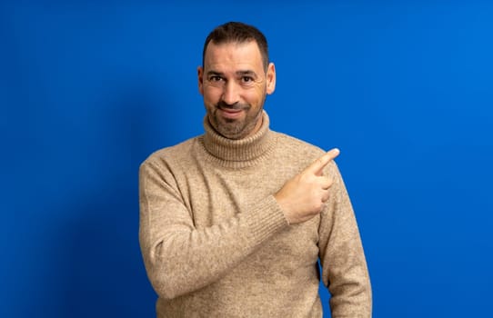 Friendly bearded Hispanic man in his 40s wearing a beige turtleneck pointing to the side, isolated over a blue background.