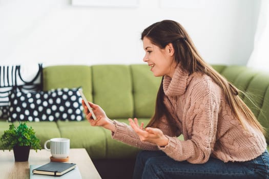 Young caucasian woman with long brown hair in pastel pink sweater and jeans working from home on her laptop and phone. Cheerful woman student studying at home.