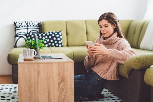 Young caucasian woman with long brown hair in pastel pink sweater and jeans working from home on her laptop and phone. Cheerful woman student studying at home.