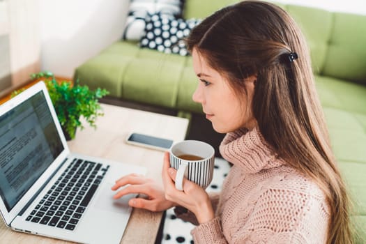 Young caucasian woman with long brown hair in pastel pink sweater and jeans working from home on her laptop and phone. Cheerful woman student studying at home.