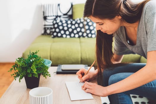 Young caucasian woman with long brown hair in jeans working from home, writing notes. Cheerful woman student studying at home.