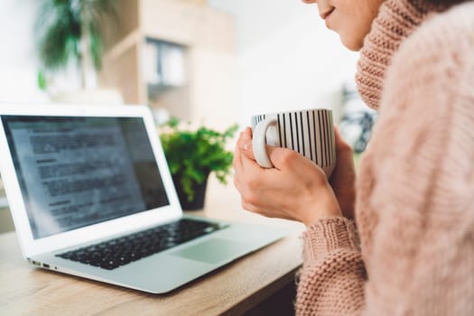 Young caucasian woman with long brown hair in pastel pink sweater and jeans working from home on her laptop and phone. Cheerful woman student studying at home.