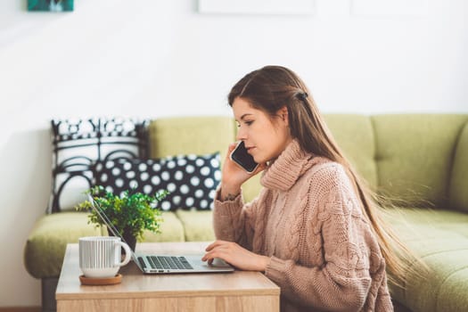 Young caucasian woman with long brown hair in pastel pink sweater and jeans working from home on her laptop and phone. Cheerful woman student studying at home.